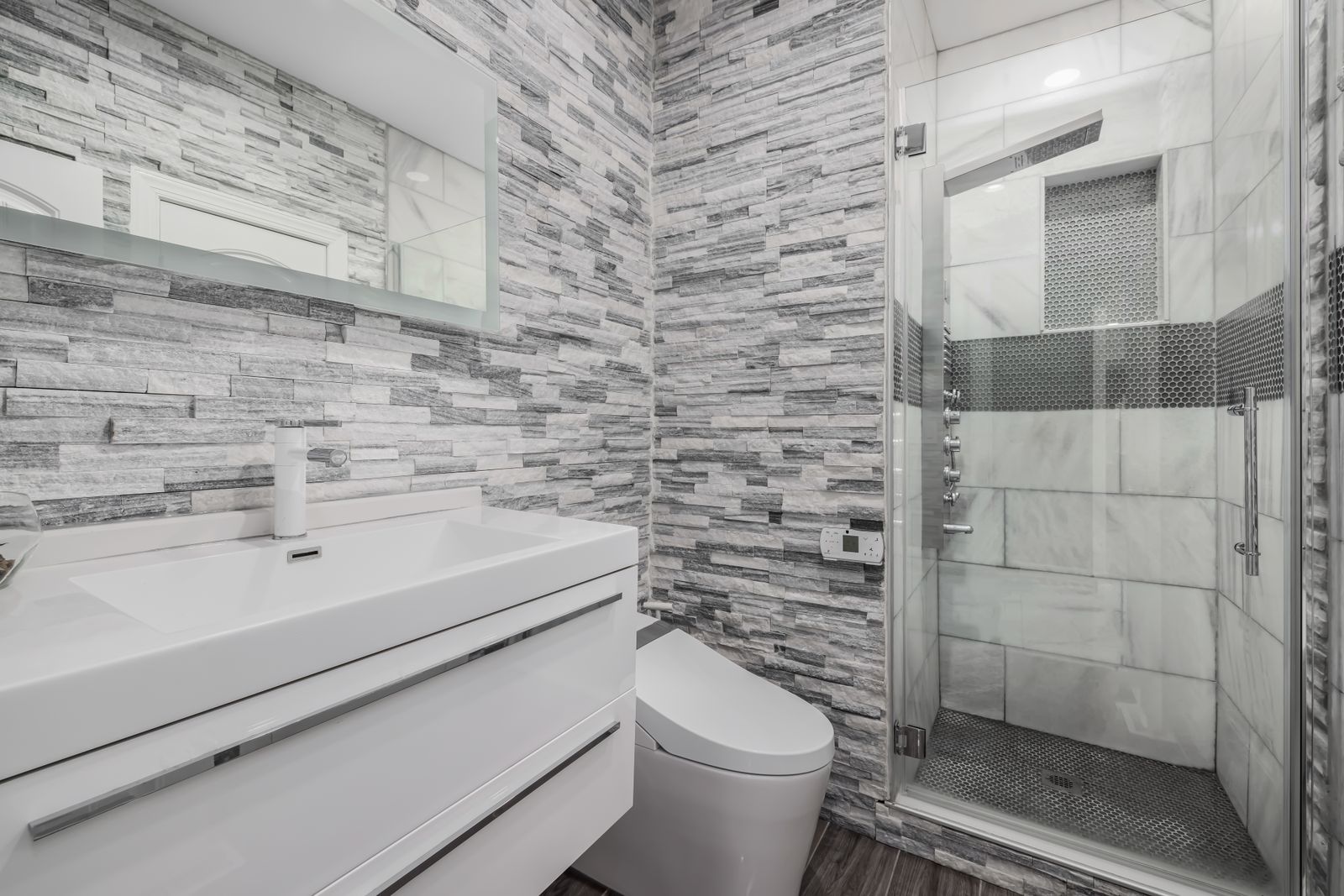 A finished modern bathroom with grey stacked-stone feature tiling, a wall-hung white vanity and wide basin with a chrome tap, a frameless glass shower with marble-look tiling, a white toilet and wood-look floor.