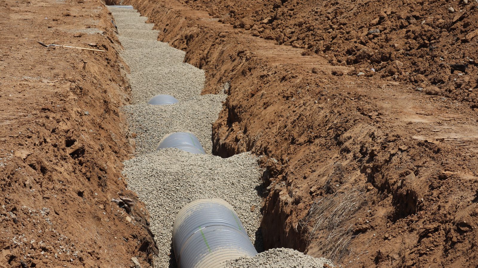 A large corrugated pipe laid in a gravel-bedded trench dug through reddish soil on a groundworks site, mid-excavation.
