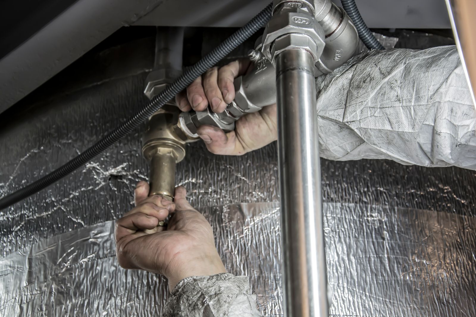 Two bare hands tightening a chrome-and-brass threaded pipe union on a bright stainless steel pipe beneath a foil-lagged water cylinder, with corrugated conduit alongside.