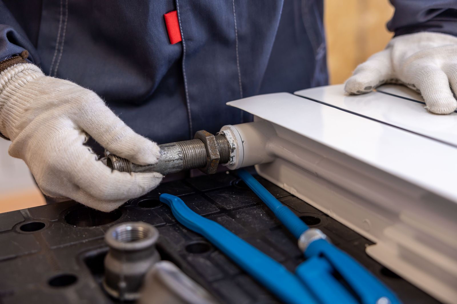 A worker in dark blue overalls and white work gloves fitting a threaded steel valve onto the end of a white panel radiator, with a blue-handled pipe wrench and steel fittings on a work tray.