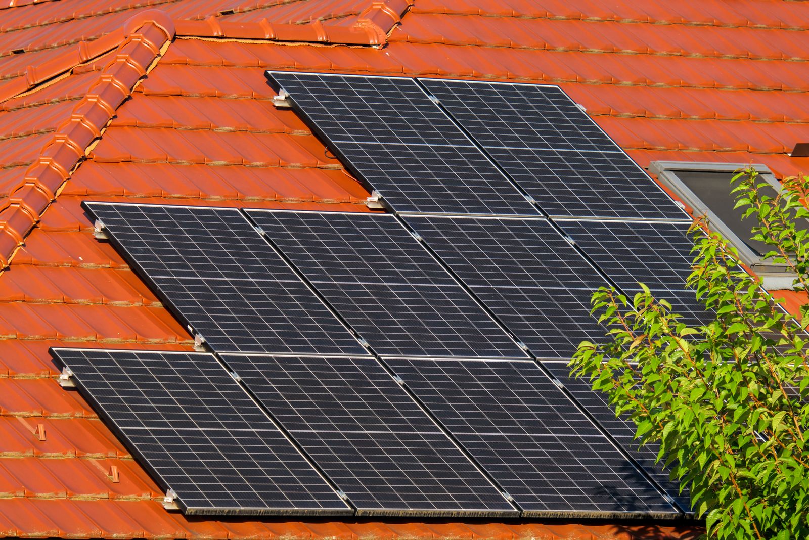 Nine dark solar panels mounted in a staggered block on a red clay-tiled pitched house roof in bright sun, with green tree foliage to the right.